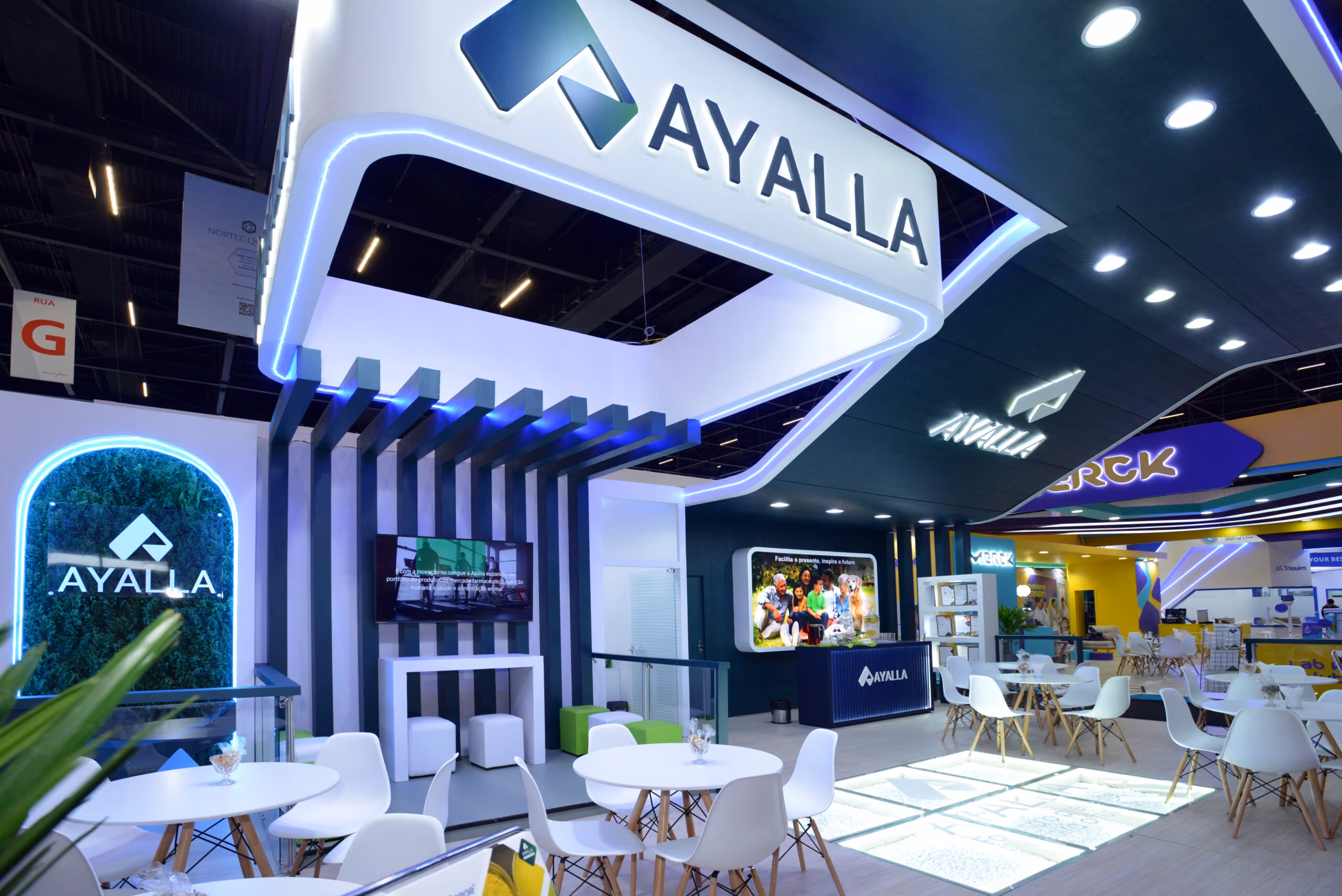 Estande moderno da Ayalla com mesas brancas, cadeiras, iluminação em LED e telas digitais.