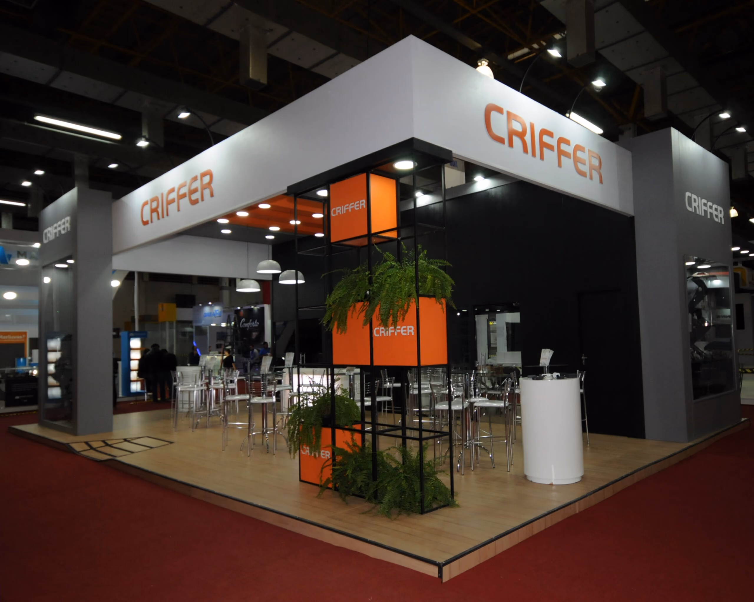 Estande da Criffer com mesas, cadeiras transparentes e plantas em ambiente de salão de eventos.